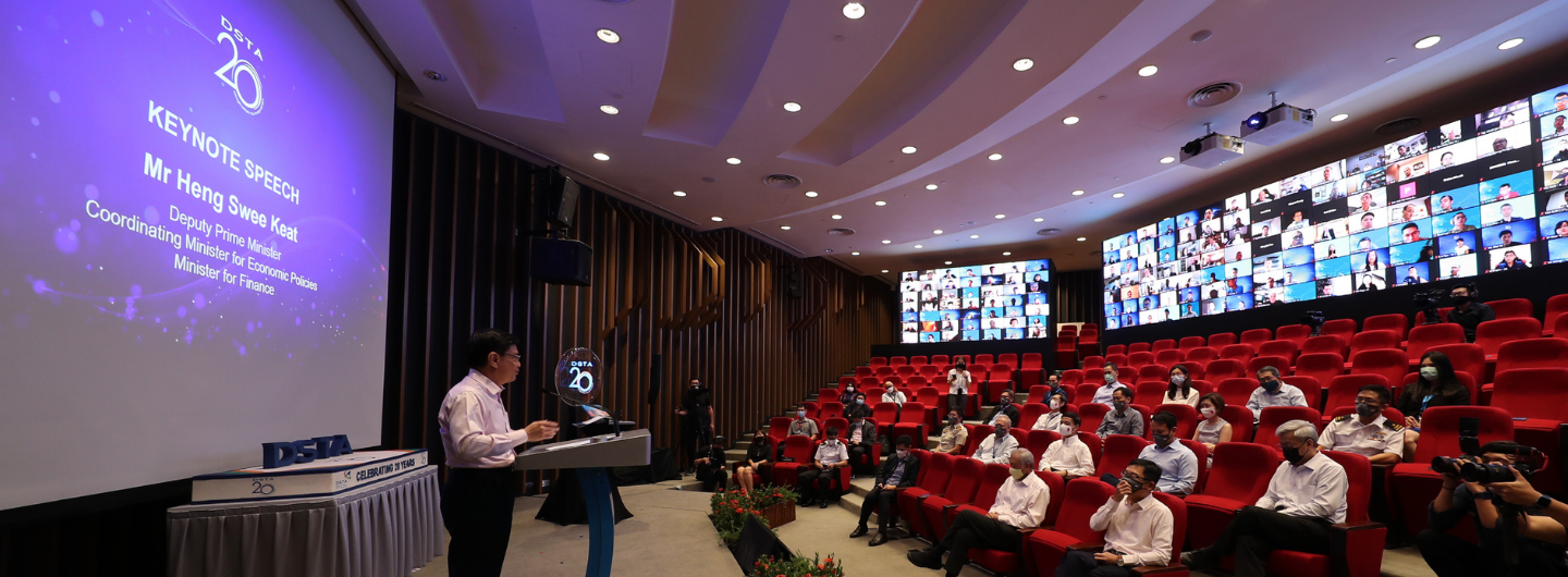 Auditorium with speaker at podium, "DSTA 20" logo, screen with many faces, and seated audience.